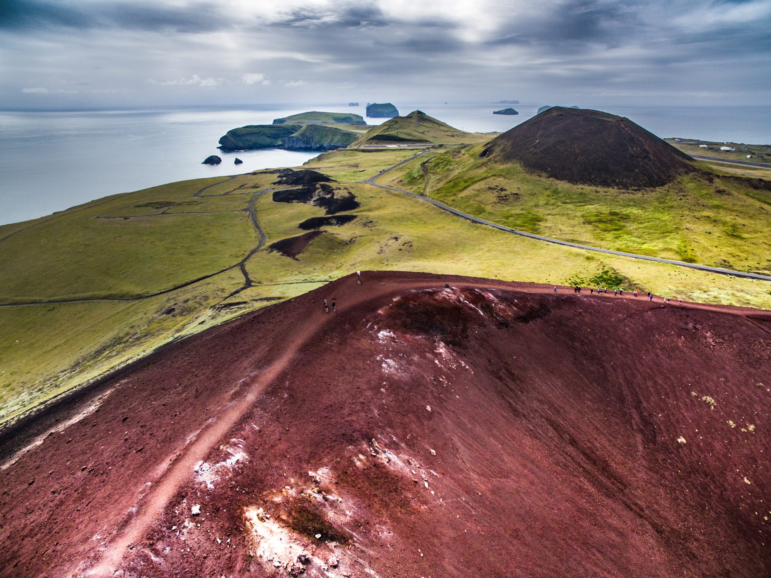 Vista aerea di un paesaggio vulcanico con terreno rosso e verde, che domina l’oceano e le isole lontane sotto un cielo nuvoloso