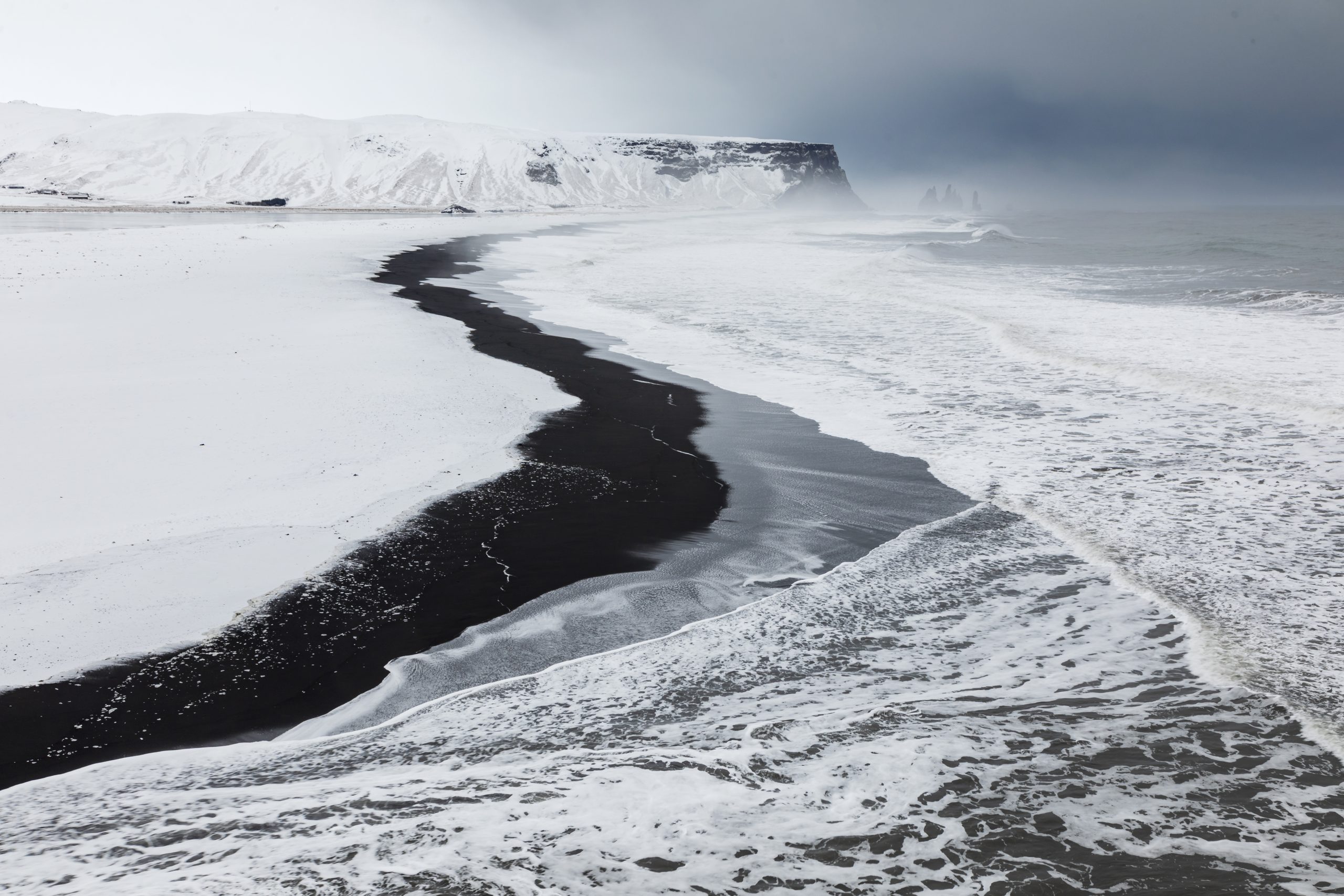 Spiaggia di sabbia nera tra scogliere innevate e onde dell’oceano